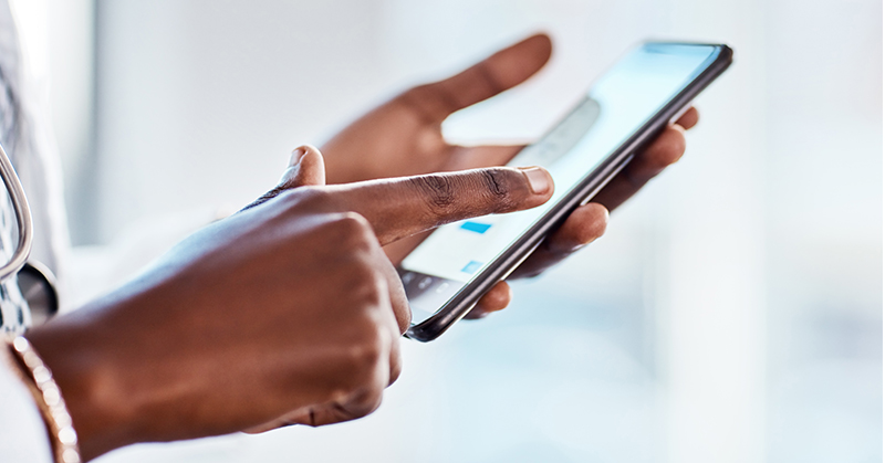 Close-up photo of the hands of a Black female health care worker typing on a cell phone in front of a blurred background. A white coat and stethoscope are partially visible on the left of the image.