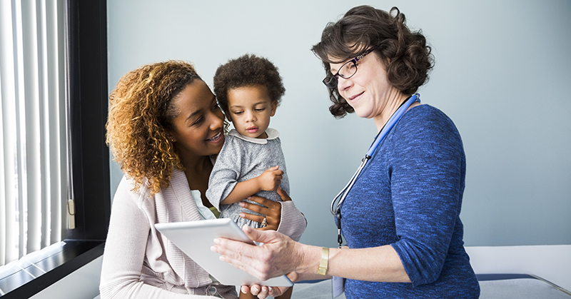 A health care provider shows a digital tablet to a mother holding a child.