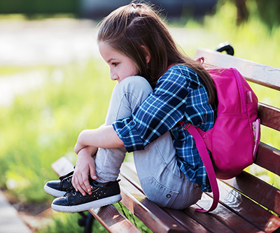 A young girl wears a backpack while sitting on a park bench, with her knees tucked under her chin.