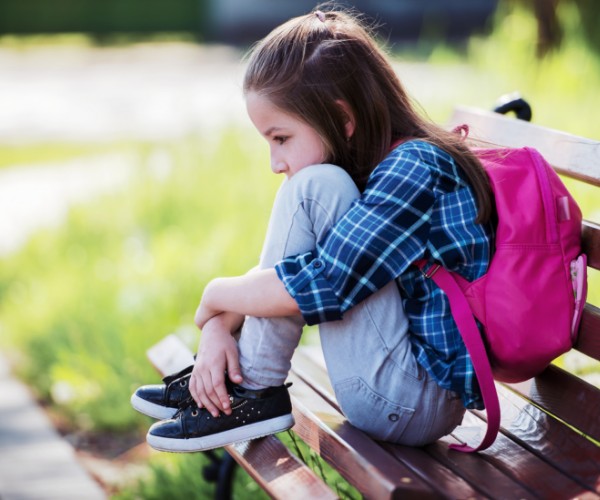 A young girl wears a backpack while sitting on a park bench, with her knees tucked under her chin.