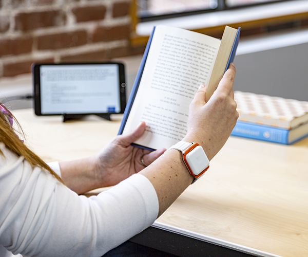 A person wears a Biosensics wrist sensor while reading a hardcover book. The sensor is a white rectangle positioned like a wristwatch.