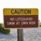 A sign reading “Caution, no lifeguard, swim at own risk” appears in the foreground. A sandy beach with dune grasses appears blurred in the background.