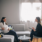 Pregnant woman, seated on couch, talking to mental health care professional with note pad and pen.