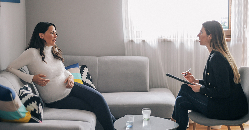 Pregnant woman, seated on couch, talking to mental health care professional with note pad and pen.
