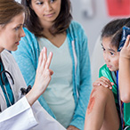 Health care worker holds up three fingers before child holding ice pack to head while parent looks on.