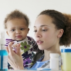 A woman holding a baby reads the label on a bottle of prescription medicine. Other medicine cabinet items appear blurred in the foreground.