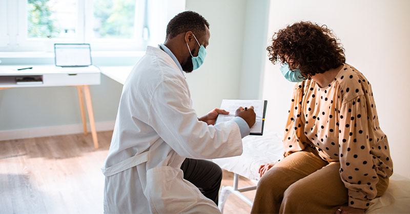 Masked health care provider in white lab coat writes on clipboard while masked patient seated on examination table looks on.