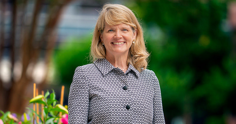 A smiling woman with trees and flowers in the background.