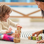 A young girl wearing a prosthetic leg sits on the floor stacking blocks with her physical therapist.