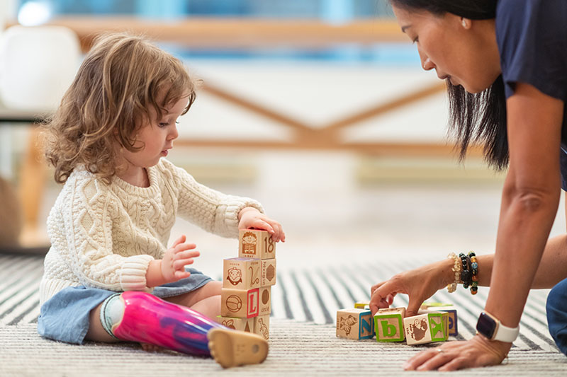 A young girl wearing a prosthetic leg sits on the floor stacking blocks with her physical therapist.