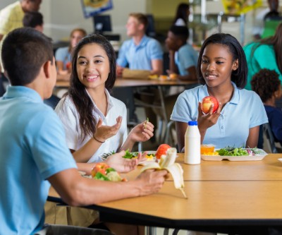 A diverse group of male and female teenagers eating lunch in a school cafeteria. Plates full of vegetables and fruit are on the tables in front of them.