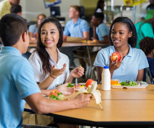 A diverse group of male and female teenagers eating lunch in a school cafeteria. Plates full of vegetables and fruit are on the tables in front of them.