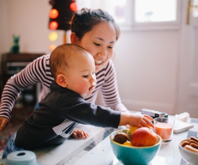 A young Asian mother in a striped shirt at a counter with her baby. The baby is reaching for a piece of fruit in a bowl on the countertop.