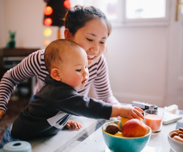 A young Asian mother in a striped shirt at a counter with her baby. The baby is reaching for a piece of fruit in a bowl on the countertop.