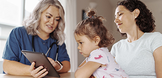 A toddler sitting on her mother’s lap gazes at a tablet device held by a healthcare provider.