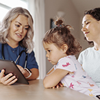 A toddler sitting on her mother’s lap gazes at a tablet device held by a health care provider.