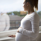 Pregnant woman looks out the window holding a hand on her belly.