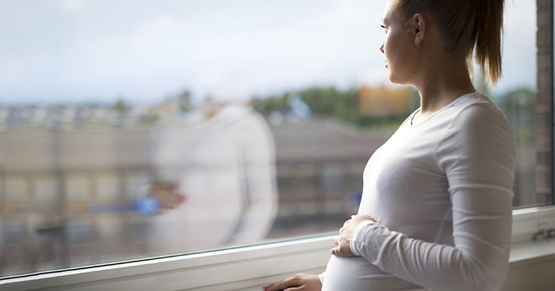 Pregnant woman looks out the window holding a hand on her belly.