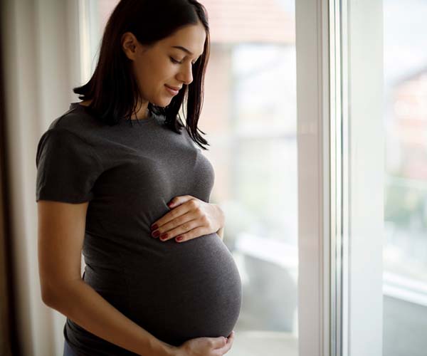 A pregnant woman stands next to a large window and looks down while holding her belly.