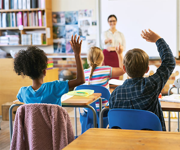 A back view of a group of children in a classroom who all have their hands raised while looking at a bespectacled teacher, who is standing at the front of the class. 