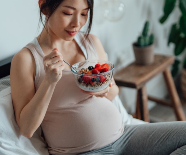 Seated pregnant woman eating a bowl of yogurt with granola and mixed berries.