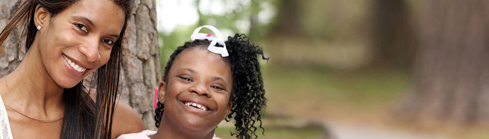A woman has her arm around a young girl with Down syndrome. Both are smiling. Trees are in the background. 