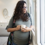 Pregnant person holding a cup, looking out a window.