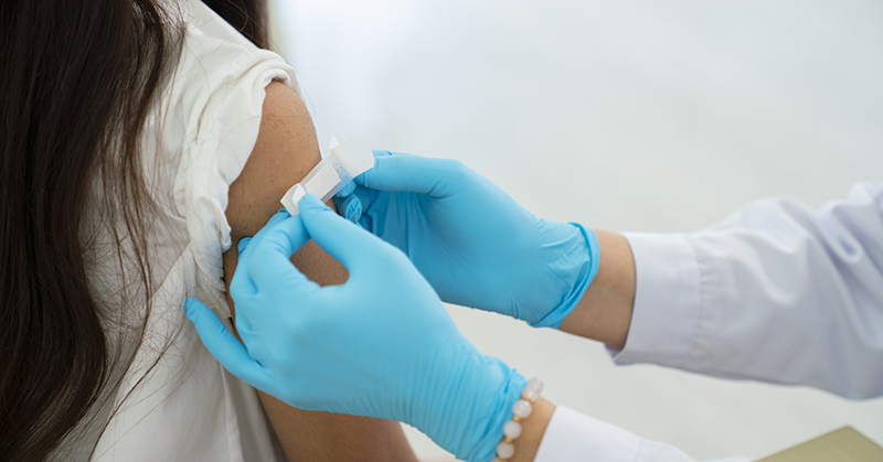 Gloved hands applying a small bandage to someone’s upper arm.