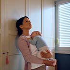 Parent holding sleeping child, leaning against a wall.