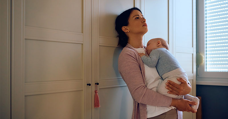 Parent holding sleeping child, leaning against a wall.