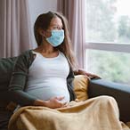 Masked pregnant woman seated in easy chair looking out window.
