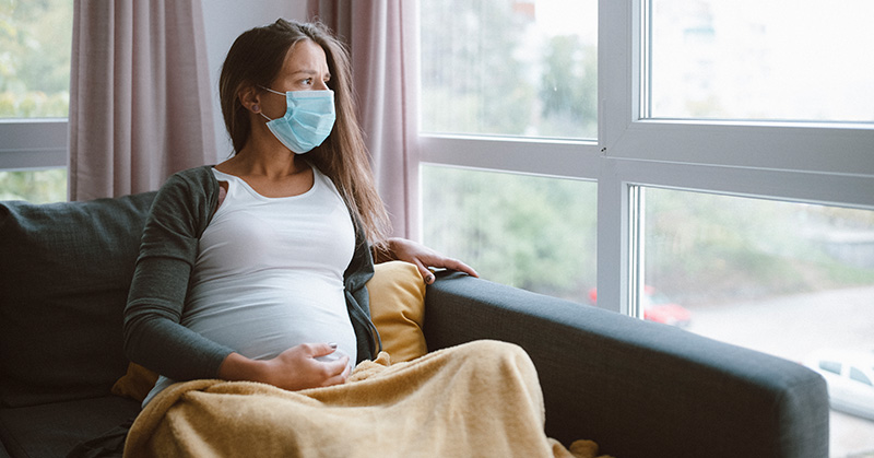 Masked pregnant woman seated in easy chair looking out window.