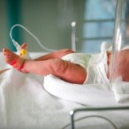An infant lying in an incubator. The infant is wearing a diaper, two medical bracelets, and a foot monitor.