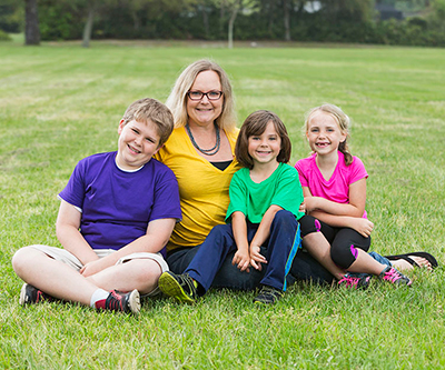 An adult and three children sit together in a grassy field, smiling and facing forward in a posed position.