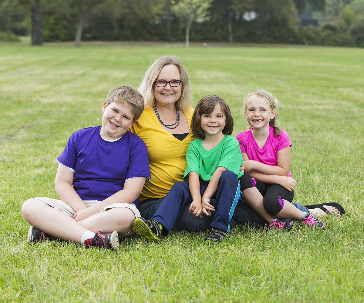 An adult and three children sit together in a grassy field, smiling and facing forward in a posed position.  
