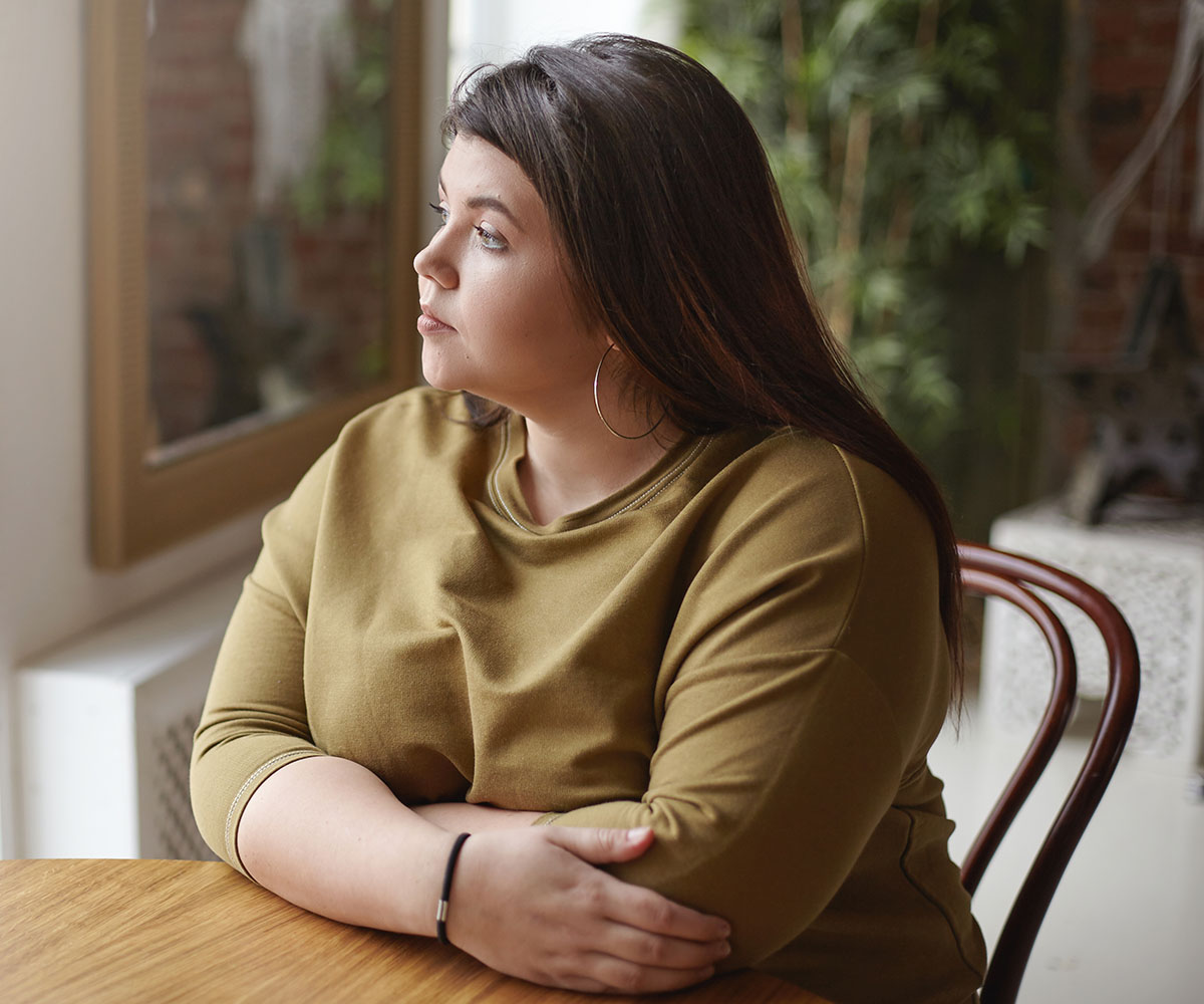 A young woman with overweight sitting at a table and looking through a window.