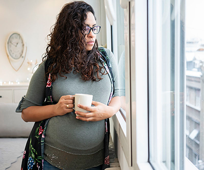 A young woman with a sad expression looks outside while holding a mug.