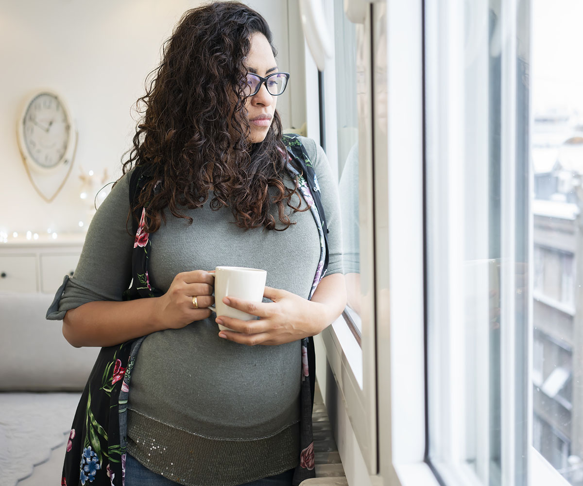 A young woman with a sad expression looks outside while holding a mug. 