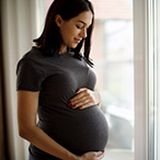 Pregnant woman with her hands on her belly, standing in front of a window.