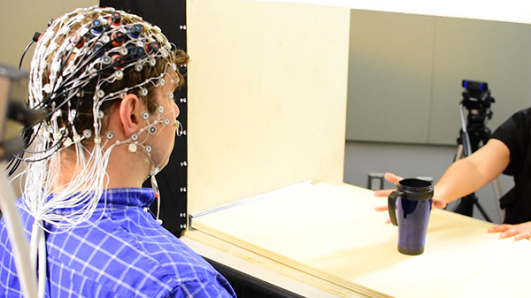 Doug is wearing the fNIRS device, which looks like a fishnet cap with sensors and wires. He is sitting across from Helga, and there is an object, a coffee thermos, between them on the table.