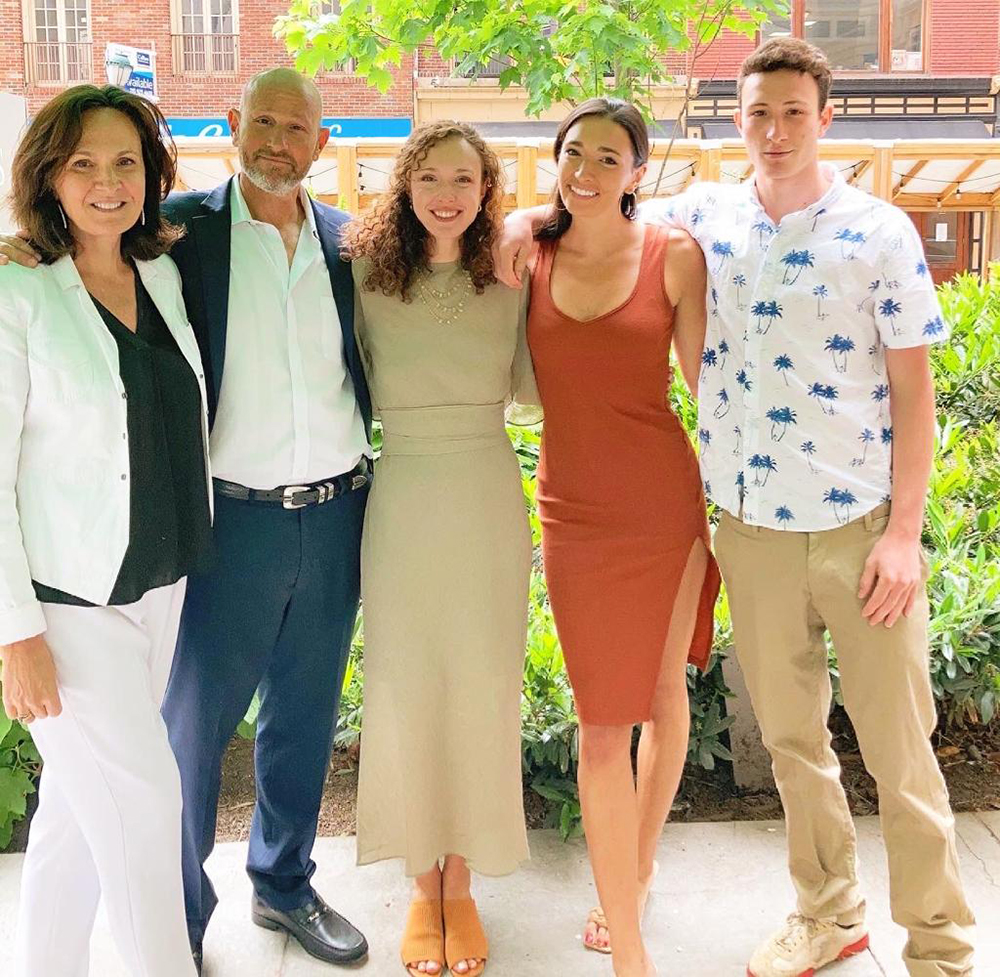 A family of five stands with their arms around each other looking at the camera. Greenery and a brick building are visible in the background.