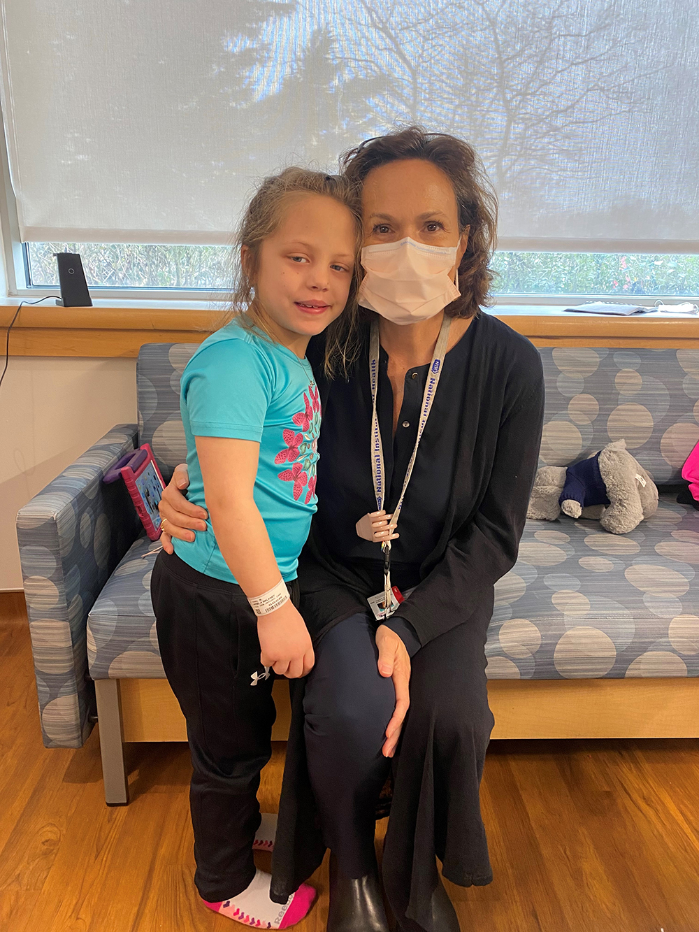 A young girl in a blue T-shirt and black pants hugs a seated woman wearing black clothes and a peach face mask.