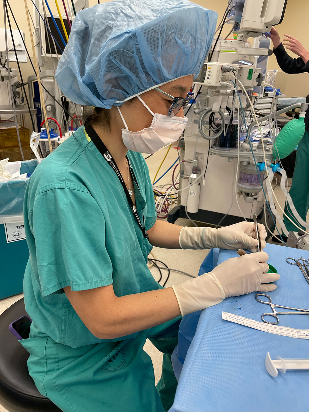 A woman wearing green scrubs, a hairnet, glasses, a facemask, and latex gloves sits at a small table working with metal tools in a small plastic dish. Medical equipment and tubing is visible in the background.