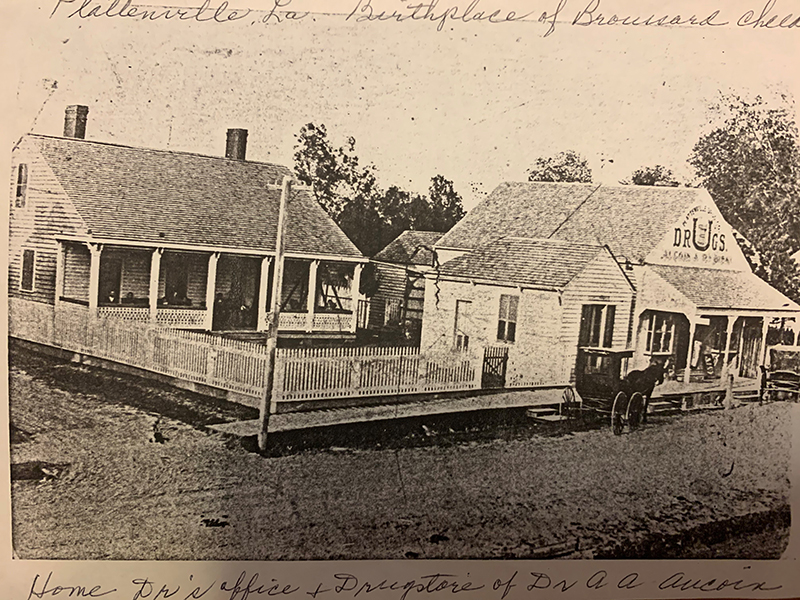 A black-and-white scan of a photograph of the old buildings. There is a horse and wagon in front of the drug store. In cursive handwriting, the following text, which is cut off at the end, is visible above the photo, “Plattensville, LA. Birthplace of Broussard Cheea.” And the following handwritten script is visible below the photo, “Home Dr’s office + Drugstore of Dr. A A Aucoin.”