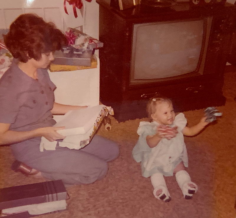 Dr. Cernich is opening a present with an adult. She is smiling and clutching two ribbons as she sits with her casts on her legs.