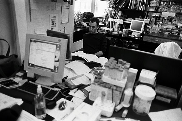 Black-and-white photo showing a desk covered with papers, test tubes, and a computer in the foreground. A man sits facing a computer screen with books, scientific research equipment, and an additional computer workstation behind him.