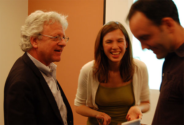 A smiling older man, a laughing woman, and a smiling younger man stand in front of a neutral background. 