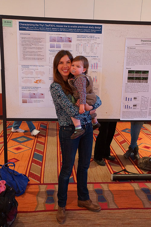 A woman holding a small toddler in her arms stands in front of a scientific research poster.