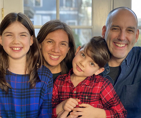 A little girl in a blue plaid top, a woman, a little boy in a red plaid shirt, and a man in a denim shirt smile in front of a window.