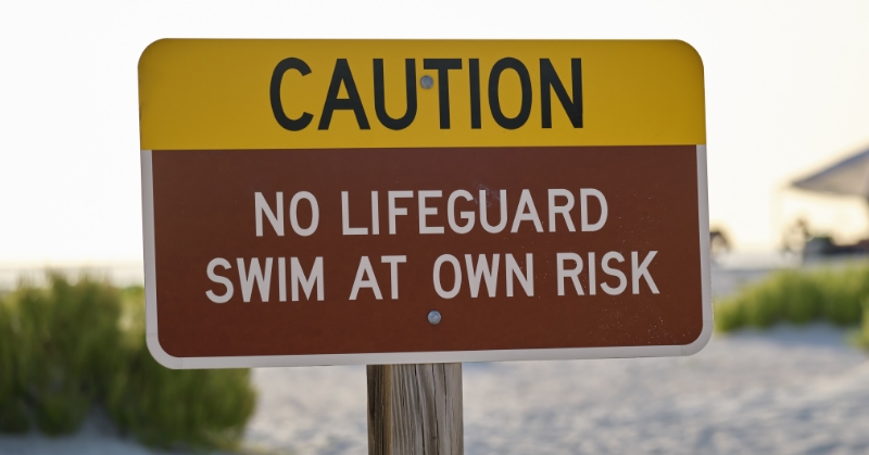 A sign reading “Caution, no lifeguard, swim at own risk” appears inthe foreground. A sandy beach with dune grasses appears blurred inthe background.